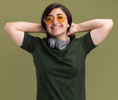 smiling young pretty caucasian girl in sun glasses and with headphones around neck puts hands on neck behind isolated on olive green background with copy space