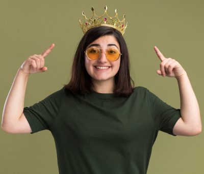 smiling young pretty caucasian girl in sun glasses with crown pointing up with two hands isolated on olive green background with copy space
